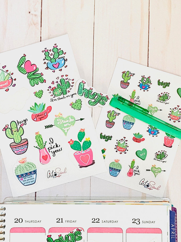 Two sheets of Cactus Valentine Stickers on a wooden background.  There is a green pen with a sticker on the tip and a planner with a single sticker also.